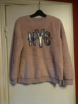 Ljuslila fluffig tröja med paljetter(barn)  - Supermjuk och fluffig ljuslila tröja med stora paljetter som bildar texten 'NYC' på bröstet. Tröjan har rund hals, långa ärmar och ribbade muddar vid ärmslut och nederkant. Perfekt för dig som gillar cozy vibes och vill sticka ut lite extra.