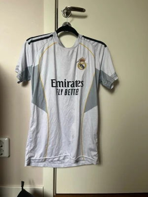 Real Madrid Mbappé matchtröja - Säljer en vit Real Madrid matchtröja med Mbappé och nummer 10 på ryggen. Tröjan har svarta och grå detaljer, klubbmärke på bröstet och sponsortryck framtill. Korta ärmar och sportig passform, perfekt för fotbollsträning eller match.