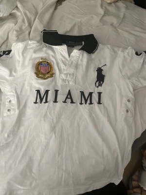 Vit Polo Ralph Lauren skjorta MIAMI - Vit skjorta från Polo Ralph Lauren med svart krage, stora broderade detaljer och texten MIAMI framtill. Klassisk pikélook med knappar vid halsen och emblem på bröstet. Perfekt för dig som gillar sportig och stilren design.