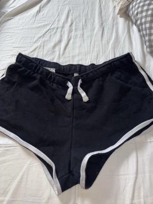 Svarta shorts med vit kant och snörning - Säljer ett par svarta shorts med vit kant och snörning i midjan. De har en sportig look och är perfekta för varma dagar. Tillverkade i mjukt material som känns skönt mot huden. Passar dig som gillar en avslappnad och enkel stil.