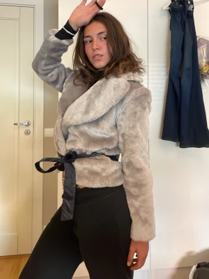 Ljusgrå pälsjacka med bälte - Snygg ljusgrå pälsjacka i fuskpäls med bred krage och svart knytbälte i midjan. Jackan har en kortare modell och ger en Rysk elegant stil. Perfekt för att hålla sig varm och bekväm på utgången