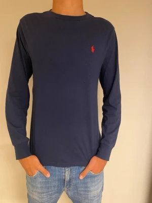 Marinblå långärmad topp Polo Ralph Lauren - Marinblå långärmad topp från Polo Ralph Lauren med klassisk rund hals och röd broderad logga på bröstet. Tillverkad i mjuk bomull som känns skön mot huden. Perfekt till jeans eller shorts för en clean och enkel stil.