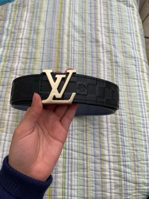 Svart rutigt Louis Vuitton bälte - Snyggt svart bälte från Louis Vuitton med klassiskt rutigt mönster och stor LV-logga i guld som spänne. Bältet är brett och tillverkat i skinn, perfekt för att lyfta vilken outfit som helst.