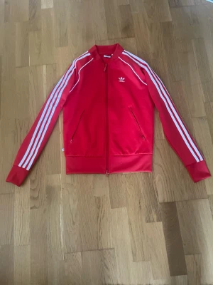 Röd Adidas track jacket med vita ränder - Snygg röd Adidas track jacket med klassiska vita ränder längs ärmarna och vit logga på bröstet. Dragkedja framtill, två fickor med dragkedja och ribbade muddar. Perfekt för en sportig och trendig look.