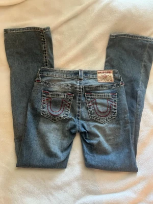 Blå jeans från True Religion - Säljer ett par blå jeans från True Religion med klassiska kontrastsömmar i vitt och rött. Jeansen har bakfickor med den ikoniska hästsko-detaljen i röd tråd. Tillverkade i denim med bälteshällor och patch bak i midjan. Modellen heter BECCA mid rise bootcut 💥 storleken är W26, Innerbenslängd: 83cm🌟 använd fåtal gånger, inga större skador ⚡️⚡️