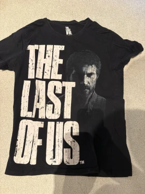 Svart The Last of Us t-shirt - Säljer en svart t-shirt med stort tryck av texten 'The Last of Us' och en bild på Joel från spelet. T-shirten har rund hals och korta ärmar. Perfekt för dig som älskar gaming och vill ha en snygg statement-tee.