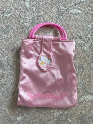 Hello Kitty rosa handväska - Söt rosa handväska i glansigt tyg med Hello Kitty-motiv framtill och texten 'Hello Kitty' i rosa. Väskan har runda rosa handtag i plast och stängs med kardborreband. Perfekt accessoar för dig som älskar kawaii och söta detaljer.