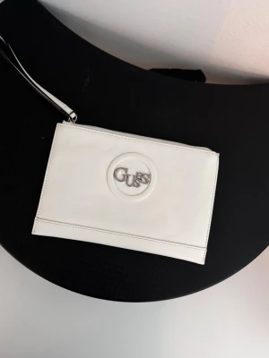 Vit kuvertväska från Guess - Snygg vit kuvertväska från Guess med silverfärgad logga framtill. Väskan har en stilren rektangulär form och handledsrem i matchande färg. Perfekt för att bära det viktigaste och ger en clean look till din outfit.