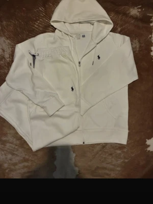 Vit ralph lauren tracksuit - Säljer en stilren vit tracksuit från Polo Ralph Lauren med dragkedja framtill och klassisk huva. Hoodien har två fickor och den ikoniska mörkblå loggan broderad på bröstet. Tillverkad i mjukt material som känns skönt mot huden. Perfekt för en avslappnad look. !PRIS KAN DISKUTERAS!