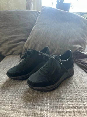 Svarta sneakers med dragkedja och snörning - Hej säljer nu ett par rieker skor i storlek 39. Inköpt för 1399kr.Dem har ej används ute bara testat på inne. Skorn har Svart mocka/nubuck på ovandelen. Sen är det en Dragkedja på sidan för enklare av- och påtagning. Den har också Grov sula med kilklack, vilket är typiskt för många Rieker modeller. Svarta lackdetaljer med krokodilpräglat mönster.