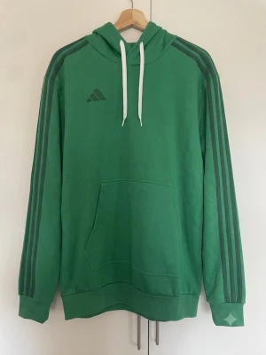 Grön hoodie med ränder från Adidas - Snygg grön hoodie från Adidas med klassiska tre ränder längs ärmarna och Adidas-logga på bröstet. Hoodien har vit dragsko i huvan och en stor magficka framtill. Tillverkad i mjukt material, perfekt för en avslappnad stil.