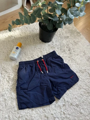 Gant badbyxor - Säljer ett par mörkblå Gant badbyxor med elastisk midja och röd dragsko framtill. Badbyxorna har fickor på sidorna och en bakficka med öljetter för vattenavrinning. Materialet är snabbtorkande syntet, perfekt för stranden eller poolen. Skriv till mig för fler frågor.🙌 12-13 år.