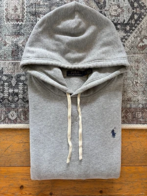 Ljusgrå Polo Ralph Lauren Hoodie - Hej! 👋 Säljer en klassisk ljusgrå Polo Ralph Lauren hoodie i storlek L med marinblå pony-logga. 🐎✨ Tröjan är i mycket gott skick, välvårdad och helt utan hål eller fläckar. Superfräsch! 100% äkta med originaltvättlappar kvar och en fullt fungerande QR-kod för enkel verifiering ✅ Det mest tidlösa och stilrena basplagget i garderoben – passar till exakt allt! Fraktar snabbt! 🚛📦💬 