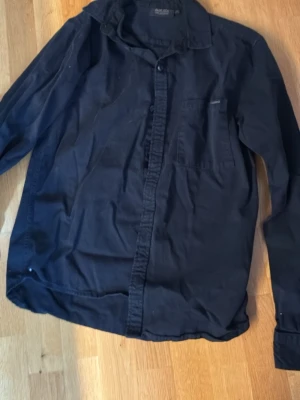 Svart skjorta från Indicode - Klassisk svart skjorta från Indicode med lång ärm och bröstficka. Skjortan har knappar framtill och är tillverkad i mjuk bomull. Perfekt för en clean och enkel stil.