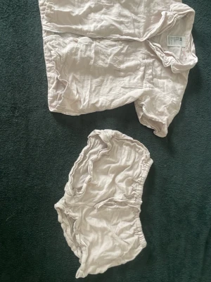 Beige pyjamasset med shorts - Säljer ett beige pyjamasset i mjukt och tunt material. Setet består av en kortärmad skjorta med knappar framtill och ett par matchande shorts med resår i midjan. Perfekt för mysiga kvällar hemma. Storlek xs