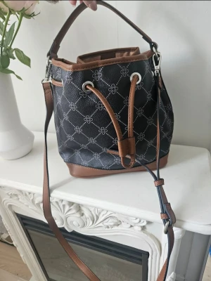 Ny bucket bag  - Snygg bucket bag axelväska i svart med brunt läderimitation och vitt mönster. Väskan har justerbar axelrem, handtag och dragskostängning. Perfekt för dig som vill ha en trendig och rymlig väska med coolt mönster och stilrena detaljer.