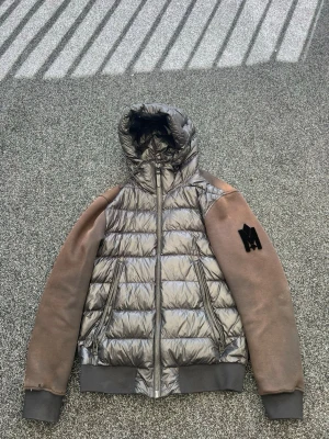 Grå cardigan - Snygg mackage pufferjacka i metallicgrå med bruna ärmar och huva. Jackan har två dragkedjefickor fram, ribbade muddar och ett svart M-märke på vänster ärm. Kroppen är quiltad och glansig medan ärmarna är i mjukare material. Köpt på NK för 7000. Pris går att förhandla.