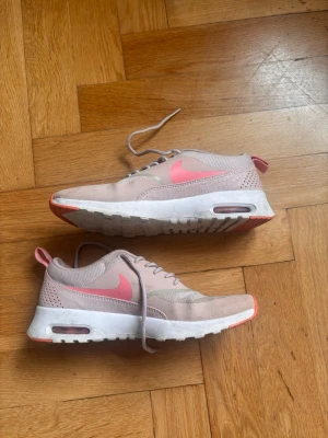 Nike rosa och vita sneakers - Snygga sneakers från Nike i ljusrosa och vitt med en rosa swoosh på sidan. Skorna har meshdetaljer för extra ventilation, synlig Air Max-dämpning i sulan och rosa detaljer på häl och yttersula. Perfekta för dig som gillar sportig och trendig stil.