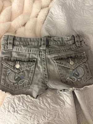 Grå slitna jeansshorts med brodyr - Säljer ett par grå jeansshorts med slitna detaljer och råa kanter. Shortsen har två bakfickor med snygg brodyr i blått och silver samt knappar på ficklocken. Klassisk femficksmodell med bälteshällor och dragkedja framtill.