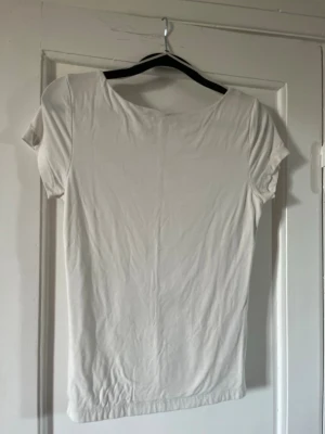 Vit topp med djup rygg - Säljer en enkel vit topp med korta ärmar och rund hals. Baksidan har en djup, rundad urringning som ger en snygg twist. Materialet känns mjukt och stretchigt, perfekt till jeans eller kjol. Passar dig som gillar minimalistisk stil.