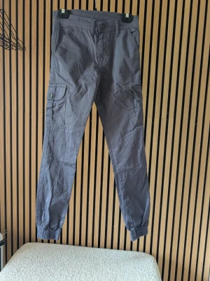 Mörkgrå cargobyxor med fickor - Snygga mörkgrå cargobyxor med resår vid benslut och flera praktiska fickor, inklusive en med knapp på sidan. Byxorna har normal passform och är tillverkade i ett mjukt bomullsliknande material. Perfekta för en avslappnad streetwear-look.