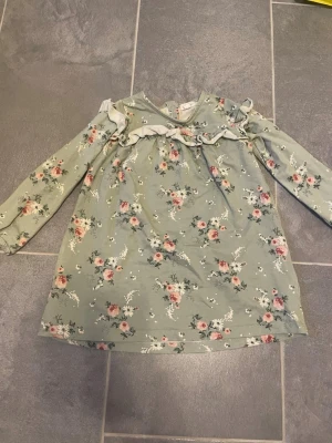 Blommig grön klänning från Newbie - Söt grön klänning från Newbie i storlek 104 med blommigt mönster i rosa och vitt. Klänningen har långa ärmar, volangdetalj över bröstet och mjukt bomullsmaterial. Perfekt för vår och sommar!