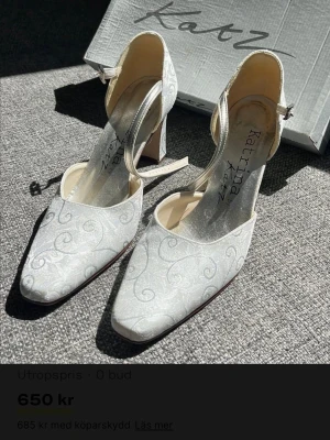 Vita pumps med mönster från Katz - Eleganta vita pumps från Katz med diskret silverfärgat, snirkligt mönster. Skorna har en klassisk spetsig tå, medelbred klack och justerbar rem runt ankeln. Materialet ser ut att vara tyg med en lätt glansig finish, perfekt för en stilren look.