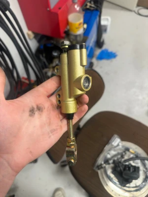 Gul bromscylinder till MC eller moped  - Säljer en guldfärgad bromscylinder i metall, troligen för bil eller motorcykel. Den har fästöglor, justerbar stång och anslutning för bromsledning. Perfekt för dig som bygger om eller behöver reservdel till bromssystem. Aldrig använd.