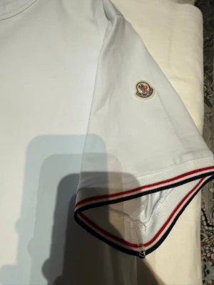Vit Moncler t-shirt med ränder - Snygg vit t-shirt från Moncler med korta ärmar och Moncler-logga på ärmen. Ärmsluten har stilrena ränder i rött, vitt och blått som ger en sportig touch. Perfekt till jeans eller shorts för en clean look.