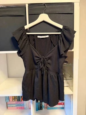 Svart blus med volang och knyt - Supersöt svart blus med volangdetaljer vid ärmarna och knyt framtill. Blusen har en v-ringad urringning. 