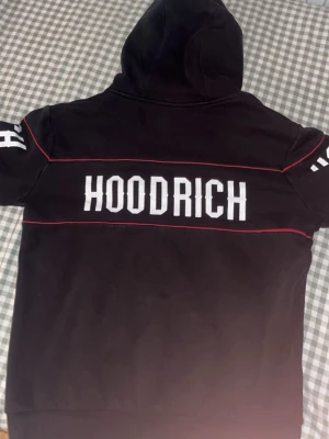 Svart hoodie från Hoodrich med tryck - Svart hoodie från Hoodrich med stor vit logga på ryggen och ärmarna. Hoodien har röda detaljer längs sömmarna och en klassisk huva med dragsko. Perfekt för dig som gillar streetwear och vill sticka ut med en fet logga.