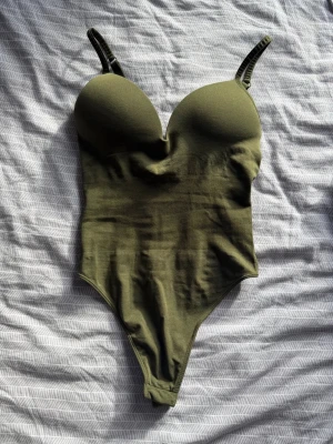 Olivgrön body från Gina Tricot - Snygg olivgrön body från Gina Tricot med vadderade kupor och justerbara axelband. Tillverkad i mjukt stretchigt material som sitter skönt mot kroppen. Perfekt att styla med jeans eller kjol för en trendig look. Endast använd en gång!