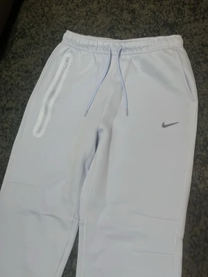 Ljusa NOCTA x Nike mjukisbyxor - Säljer ett par ljusa mjukisbyxor från Nike och NOCTA med raka ben, resår i midjan och dragsko. Byxorna har en vit dragkedjeficka med reflekterande detalj och NOCTA-logga på benet. Perfekta för en chill och sportig look.