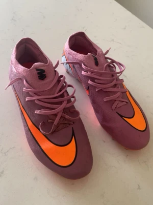 Lila Nike Mercurial fotbollsskor - Säljer ett par lila Nike Mercurial fotbollsskor med orangea detaljer och svart logga. Skorna har snörning och en strumpliknande passform för extra komfort på planen. Tillverkade i syntetmaterial som ger bra grepp och kontroll på bollen.