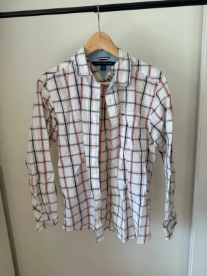 Vit rutig skjorta från Tommy Hilfiger - Vit skjorta från Tommy Hilfiger med rött och blått rutmönster. Klassisk krage, långa ärmar och knappar framtill. Tillverkad i bomull för en skön känsla och avslappnad look. Perfekt för dig som gillar stilrena och tidlösa plagg. Plagget har en liten fläck som säkert går bort med tvätt.