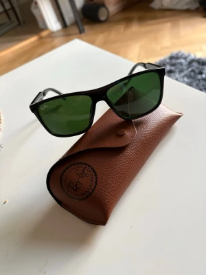 Svarta Ray-Ban solglasögon - Snygga svarta solglasögon från Ray-Ban med klassisk rektangulär form och gröna glas. Kommer med originalbrunt fodral i skinnimitation. Perfekta för soliga dagar och ger en cool look.