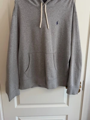 Grå hoodie från Polo Ralph Lauren - Säljer en stilren grå hoodie från Polo Ralph Lauren med klassisk blå logga på bröstet. Hoodien har vit dragsko i huvan och en stor magficka framtill. Tillverkad i mjukt bomullsmaterial som är skönt mot huden. Perfekt för en avslappnad look.