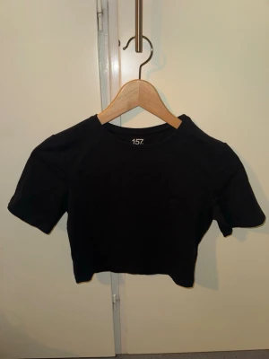 Svart cropped t-shirt från 157 - Svart cropped t-shirt från 157 med rund halsringning och korta ärmar. Tillverkad i mjuk och stretchig bomullsblandning (95% bomull, 5% elastan) som sitter snyggt och bekvämt. Perfekt till jeans eller kjol för en enkel och stilren look.