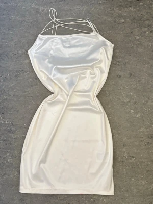 Vit satin klänning med tunna band - Snygg vit kort klänning i satin med tunna axelband som korsas över axlarna. Klänningen har en slät, glansig yta och figurnära passform. Perfekt för dig som vill ha en enkel men elegant look.