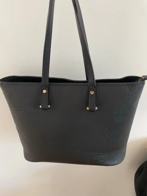 Svart shoppingväska med guldiga detaljer - Stilren svart shoppingväska i konstläder med två långa axelband och guldiga metalldetaljer. Väskan har dragkedja upptill och ett stort innerfack med extra ficka med dragkedja. Perfekt för dig som vill ha plats för allt och gillar en klassisk look.😊 Väskan har några få skavanker som man ser på bilderna, men dom syns inte tydligt när man har på sig väskan🤗