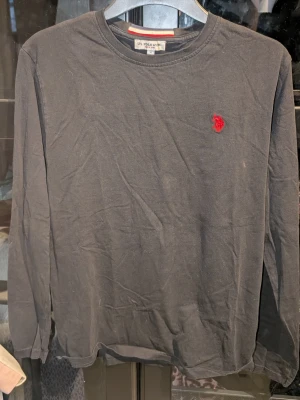 Svart långärmad tröja U.S. Polo Assn - Klassisk svart långärmad tröja från U.S. Polo Assn. med rund halsringning och röd broderad logga på bröstet. Tillverkad i mjuk bomull som känns skön mot huden. Lite urtvättad 
