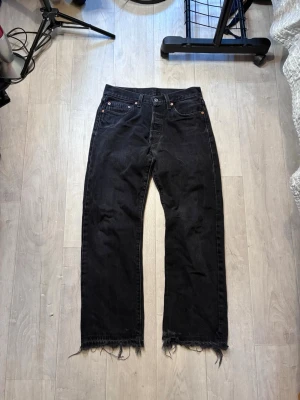 Svarta Levi’S 501  jeans  - Säljer ett par svarta Levis 501 jeans, fransig kant nedtill. Jeansen har klassisk femficksdesign och knappgylf. Materialet är kraftig denim i bomull. Perfekta för en avslappnad och trendig look.