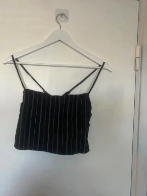 Svart randig linnetopp med smala band - Snygg svart linnetopp med vita vertikala ränder och tunna axelband. Toppen har en croppad passform och knytning i ryggen för extra detalj. Perfekt för varma dagar och enkel att styla med jeans eller kjol.