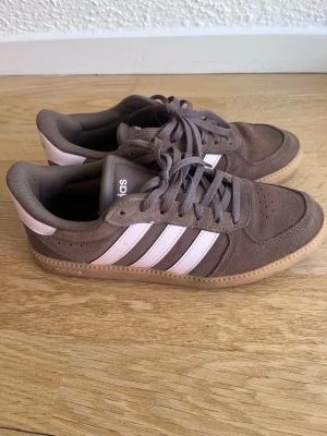 Bruna Adidas sneakers med rosa detaljer - Säljer ett par bruna Adidas sneakers i mocka med klassiska vita ränder på sidan och ljusrosa detaljer vid hälen. Skorna har snörning och en gummisula i naturfärg. Perfekt för dig som gillar en stilren och sportig vibe.