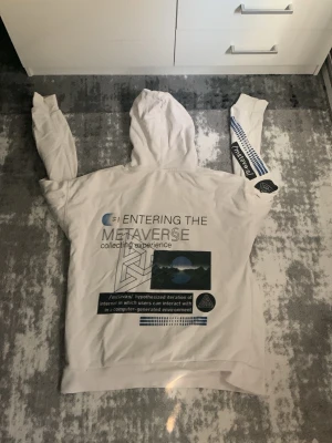 Vit hoodie med metaverse-tryck - Säljer en vit hoodie med stor huva och tryck på både fram- och baksida. På ryggen står det 'Entering the Metaverse' med futuristiska grafiska detaljer och text. Ena ärmen har också tryck. Hoodien har magficka och ribbade muddar.
