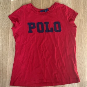 Röd t-shirt från Polo Ralph Lauren - Röd t-shirt från Polo Ralph Lauren med stor mörkblå POLO-text framtill. Klassisk rund halsringning och korta ärmar. Tillverkad i mjuk bomull som känns skön mot huden. Perfekt för en avslappnad och stilren look.