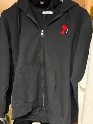 Svart zip hoodie från Ami Paris - Säljer en svart hoodie med dragkedja från Ami Paris. Den har en huva med dragsko, kängurufickor och ett rött Ami-hjärta broderat på bröstet. Perfekt för en chill och stilren look. Materialet känns mjukt och skönt mot huden.