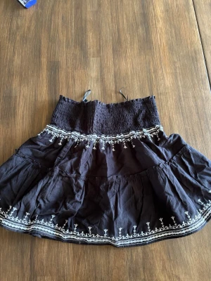 Svart kortkjol med broderi från H&M - Svart kortkjol från H&M Divided med vitt broderi längs nederkant och midja. Kjolen har smockad midja och volangig form som ger en luftig känsla. Perfekt för dig som gillar detaljer och en söt, avslappnad stil.