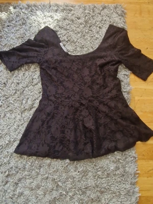 Svart spetspeplumtopp - Snygg svart peplumtopp med blommigt spetsmönster och halvlånga ärmar. Toppen har en djup rundad urringning och markerad midja som ger en feminin siluett. Perfekt att styla med jeans eller kjol för en trendig look.