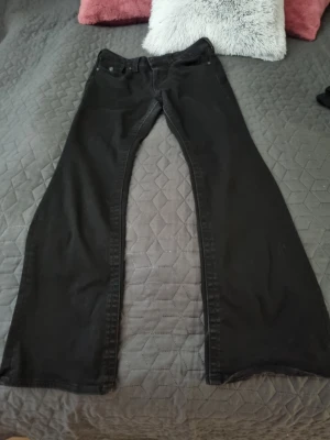 Svarta bootcut jeans True Religion - Snygga svarta jeans från True Religion med bootcut passform och klassiska detaljer. Jeansen har två bakfickor med lock och knapp samt tydliga sömmar. Perfekta för dig som gillar en avslappnad men trendig stil. Använd i fåtal gånger i storlek 30 motsvarar  42. Midjemått 42yterbenlängd 109, innerbenslängd 82.  Kolla bild två och fyra. Den är fint svart men på bilden ser ljusare ut på grund av kameran. 